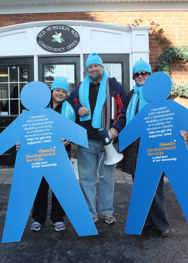 Three members holding up Developmental Services signs shaped like people. The image is outside of Ted McMeekin's MPP Constituency Office