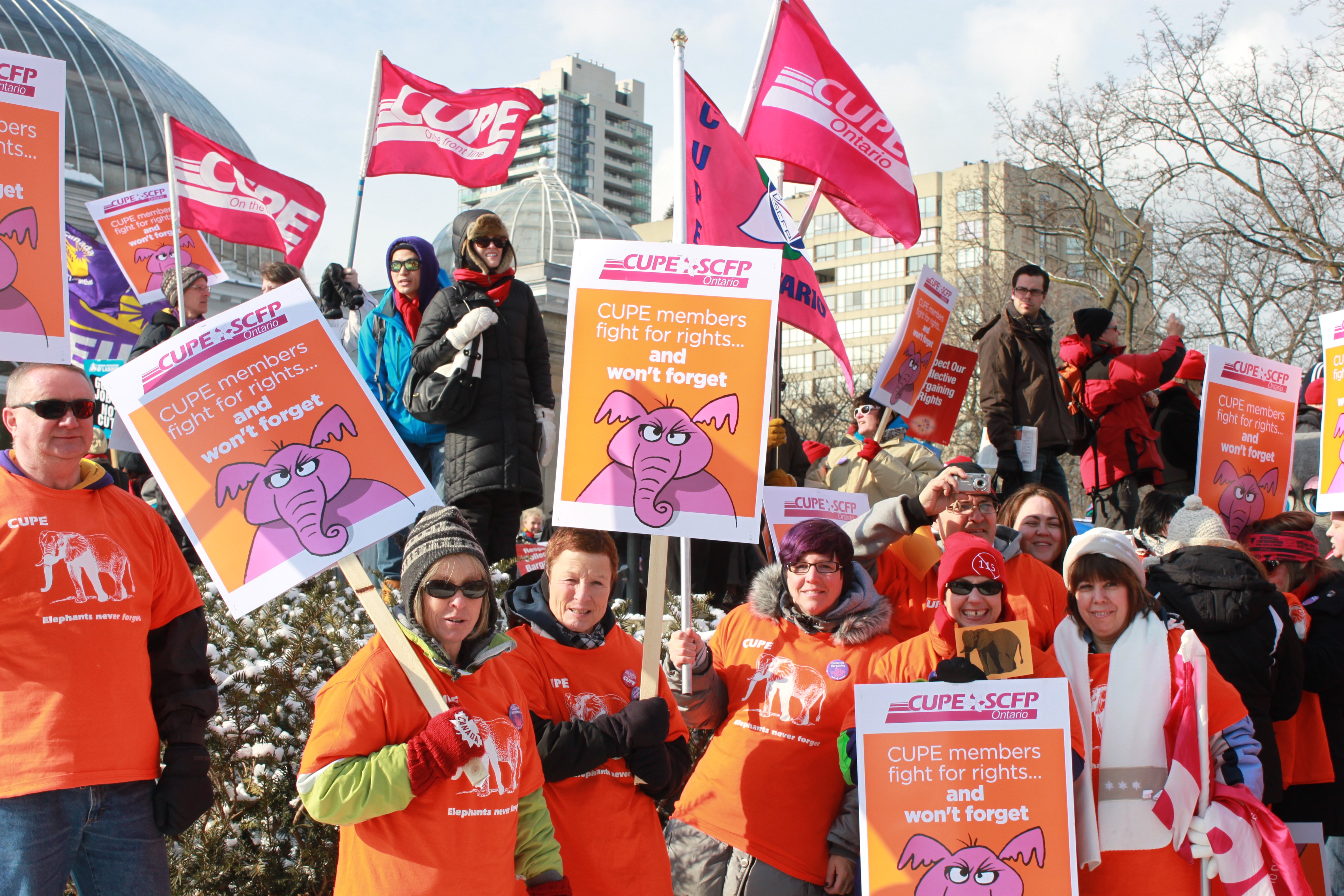 Members outside of Allan Gardens in Toronto, holding up signs that say "CUPE members fight for rights... and won't forget". The sign has a hand-drawn angry pink elephant on it