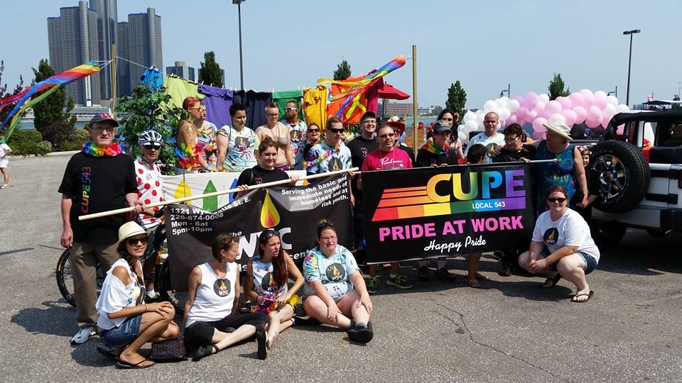 Over a dozen CUPE members pose in front of a banner at Windsor-Essex Pride. The words on the banner read 