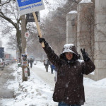 A person holding the sign "On Legal Strike" above their head in one hand, and making the "peace" sign with the other hand outside in the snow