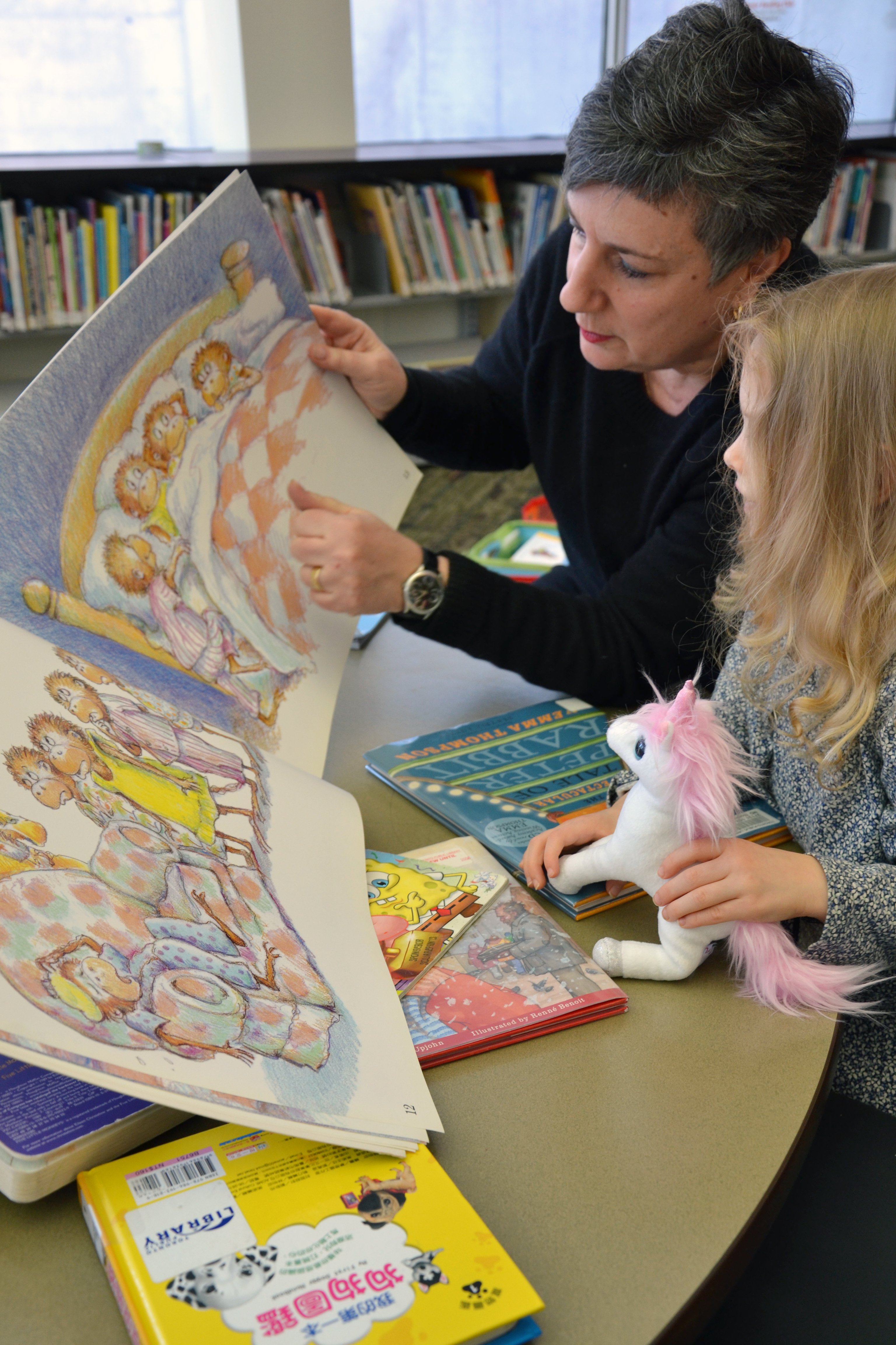 A library worker reading a book to a small child. The child is holding a pink unicorn.
