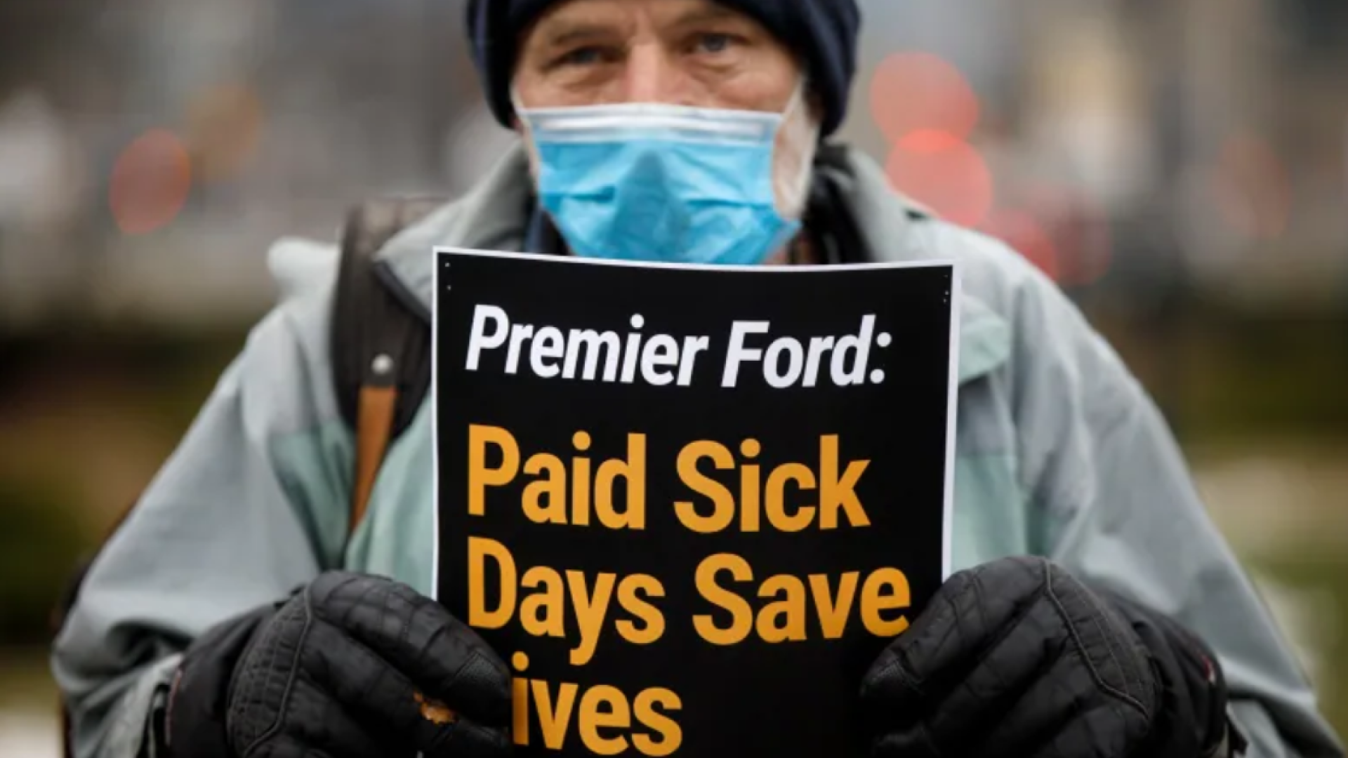 Image shows a person, outdoors, wearing a raincoat, gloves, a toque, and a face mask and holding a sign that reads 