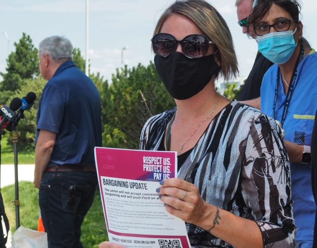 A hospital worker holds a colourful paper sign that reads "Respect Us; Protect Us; Pay Us" at an outdoor rally.