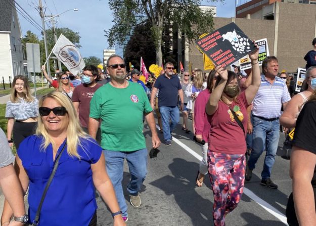 Image shows many people in colourful t-shirts marching outdoors down the middle of a street.