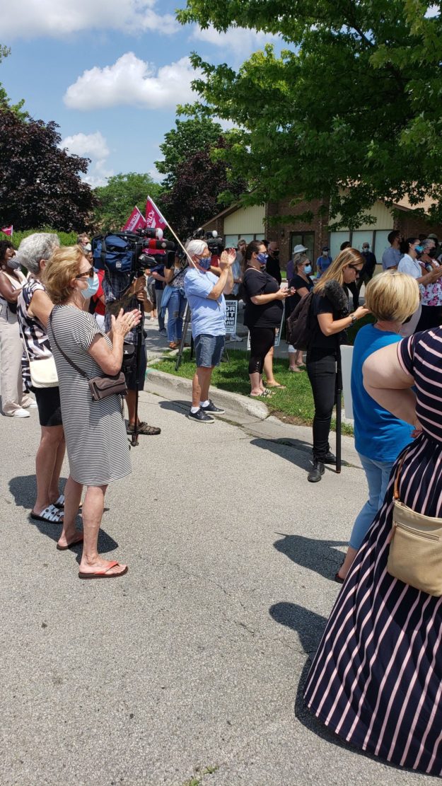 Image shows people, masked, standing around outside at Orchard Villa rally