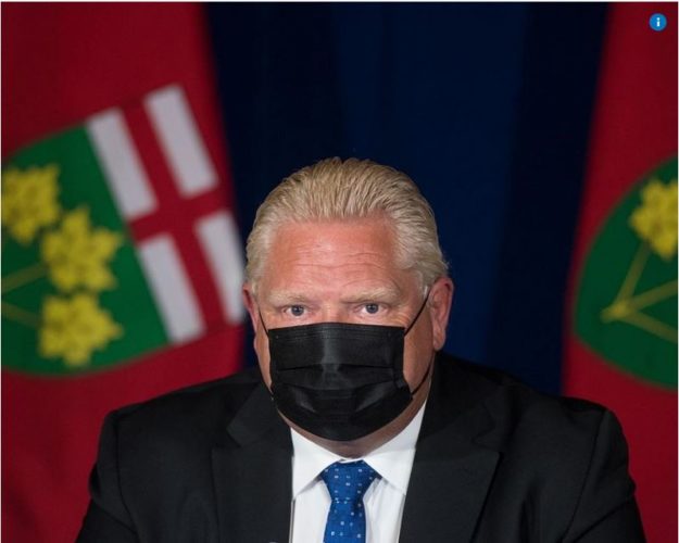 Picture shows Rob Ford, wearing a black face mask, wearing a suit and positioned in front of two hanging Ontario flags.