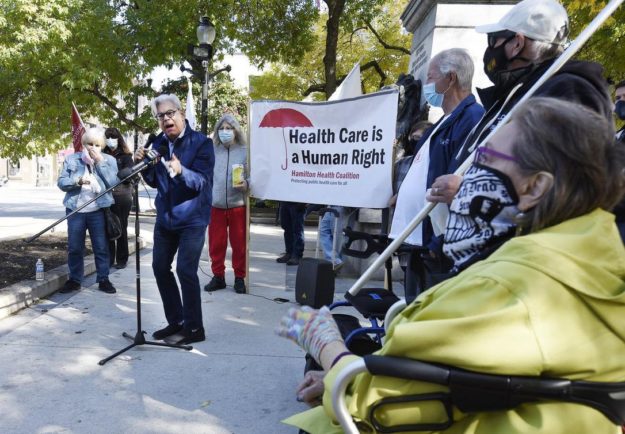 Image shows Fred Hahn, President of CUPE Ontario, standing at a microphone outdoors, with people masked around him, holding a banner that reads "Health Care Is a Human Right"