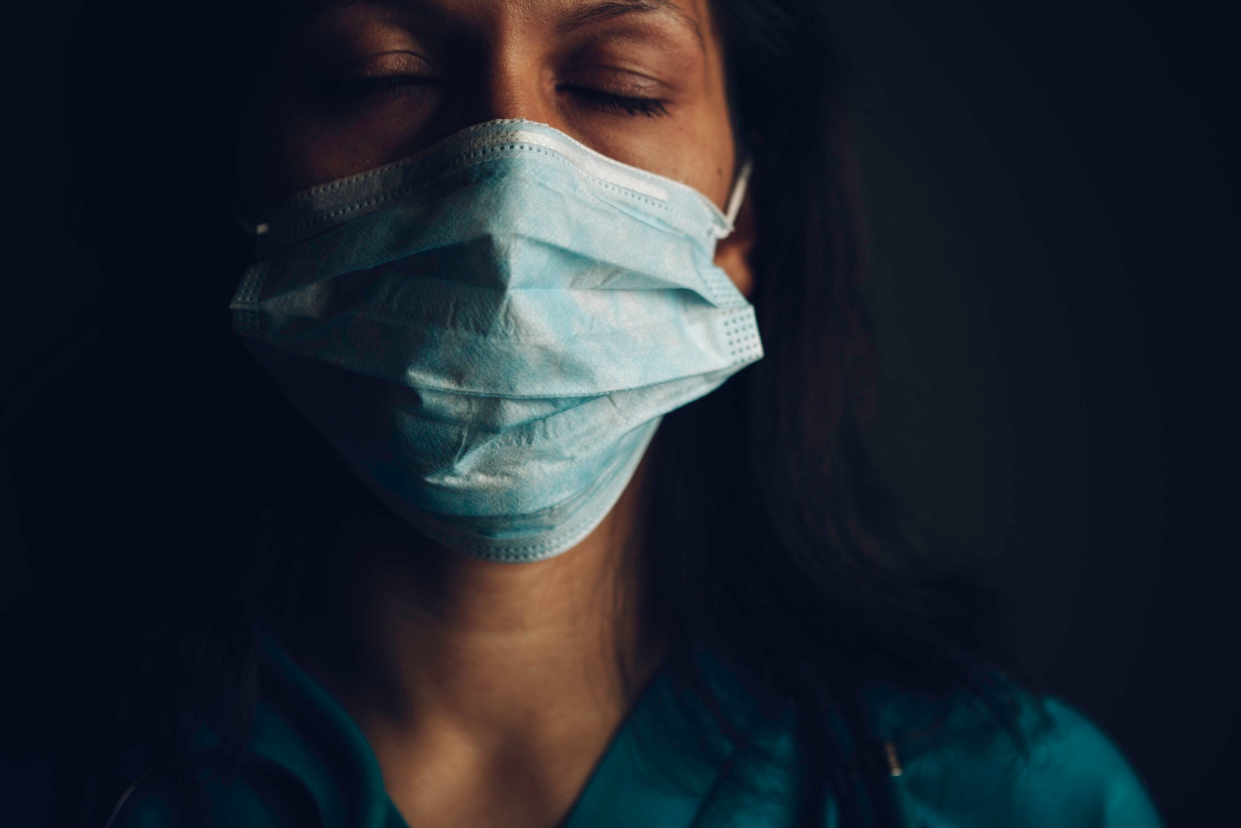 Image shows a healthcare worker's head and neck. Their eyes are closed, they're wearing a medical mask, and they're wearing scrubs.