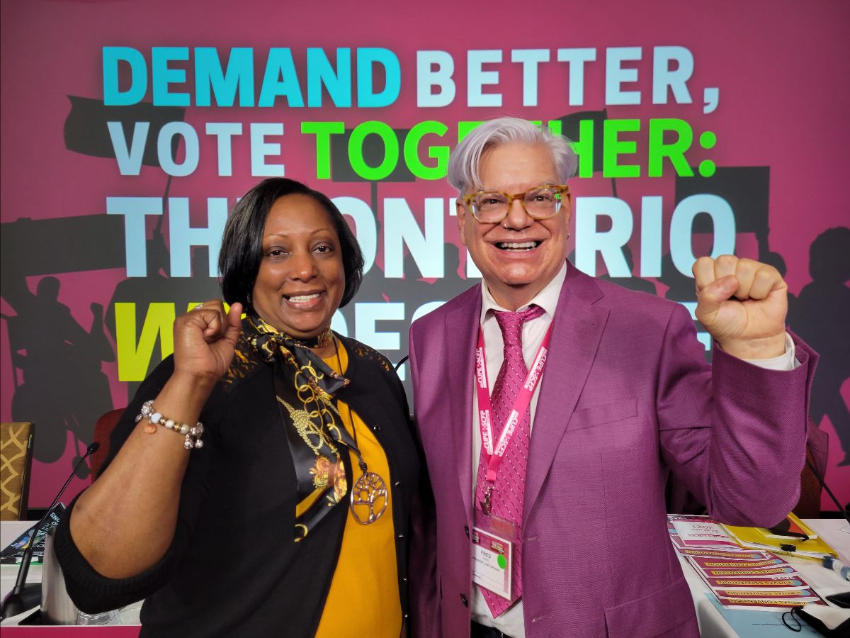 Image shows Yolanda McClean and Fred Hahn with their fists raised to indicate labour union solidarity. They stand against a backdrop of the Convention logo in English, which read 