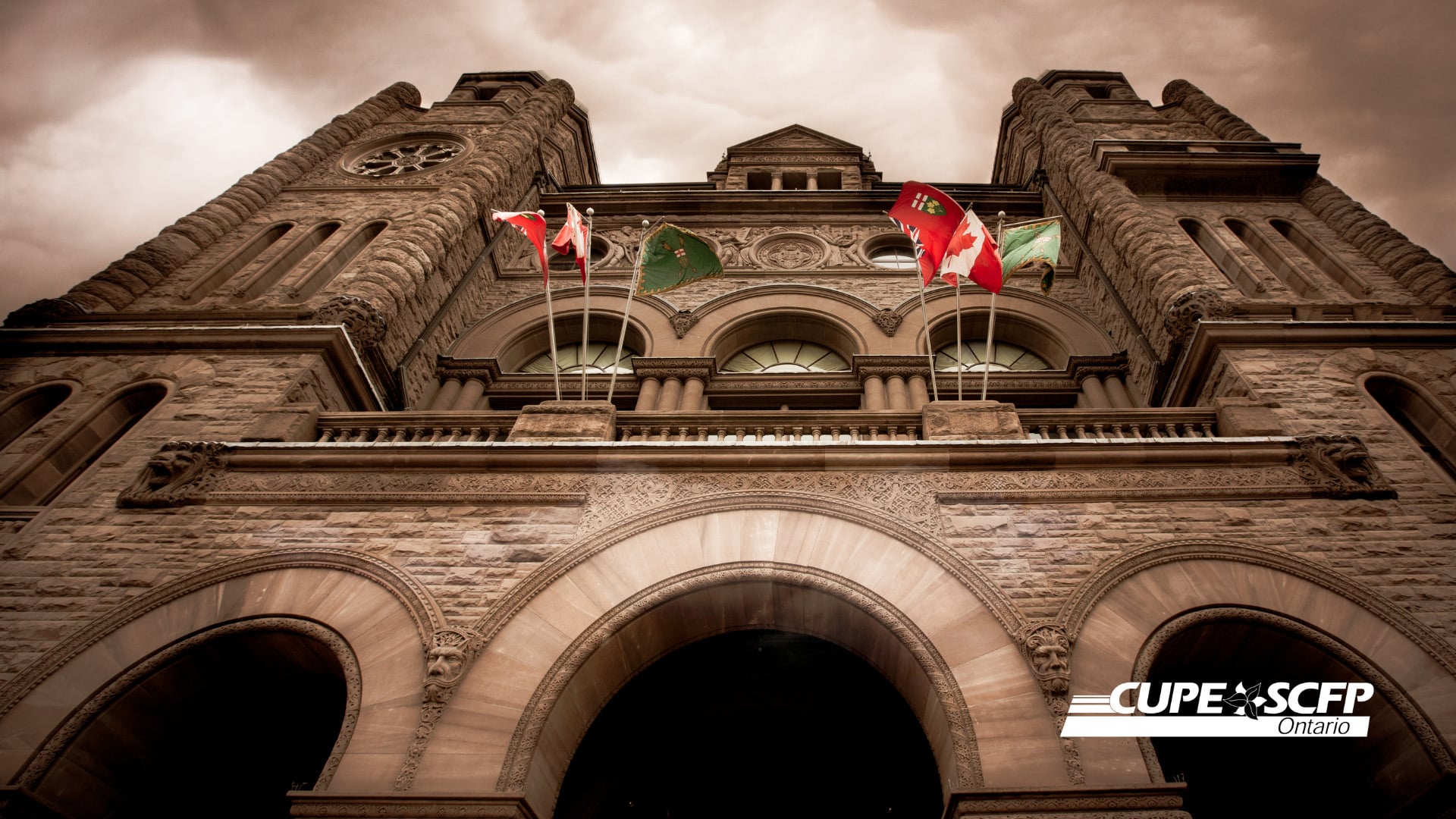 Image shows Toronto's Old City Hall from below, looking up. Canada and Ontario flags, among others, are flying on flag poles partway up the building, above large stone arches that form the entrance to the building.