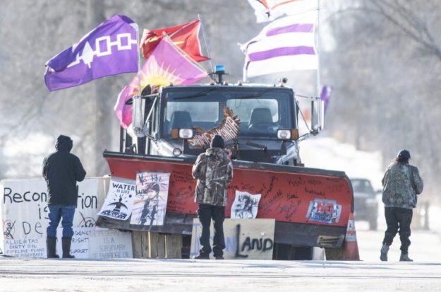 Fred-op-ed_Apr2022-2 Image shows three people dressed in winter gear looking towards and around a snow plow that has many cardboard signs and four flags on and around it, as part of the so-called "Freedom Convoy."