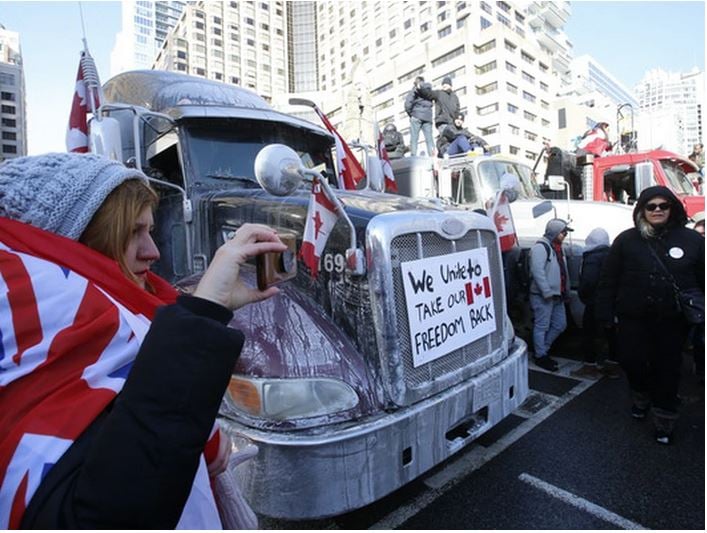 Fred-op-ed_Apr2022 Image shows people wearing Canadian flags as capes flanking a truck with a sign on the front of it that reads "