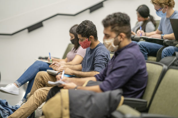 College Students wearing masks in a lecture hall Image shows university students in a lecture hall writing by hand in notebooks. All are wearing face masks.