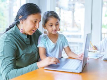 A Native American woman sits at the dining room table and works on a laptop computer as her adolescent son works on homework next to her. The woman's elementary age daughter is sitting on her lap and is looking at the computer screen.