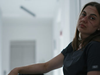 From below exhausted young doctor with bruises on face from protective glasses and masks having hot drink while relaxing on floor after hard work with patients COVID 19 infected in hallway of modern clinic