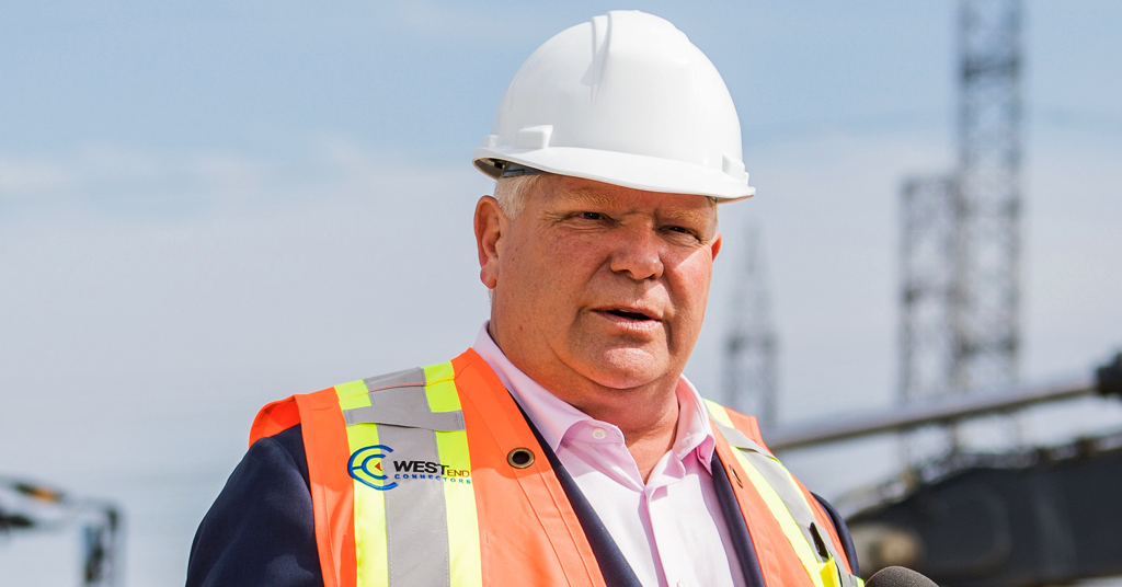 Image shows Premier Doug Ford in a hard hat and reflective vest, in front of power line towers in the background.
