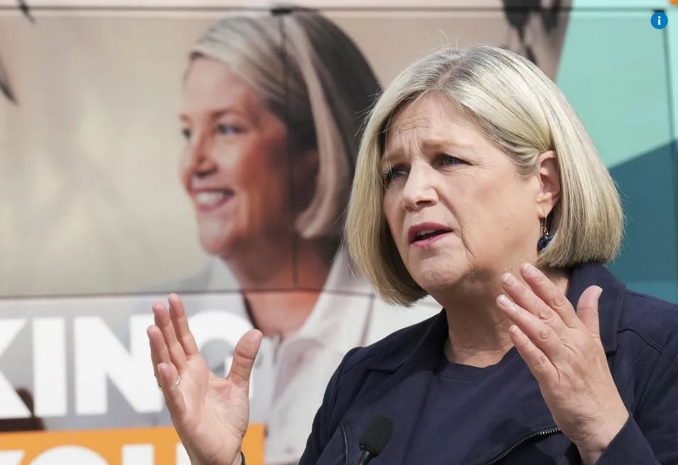 Image shows Andrea Horwath speaking, with both hands raised, with palms facing each other. She stands against a background that shows her election banner.