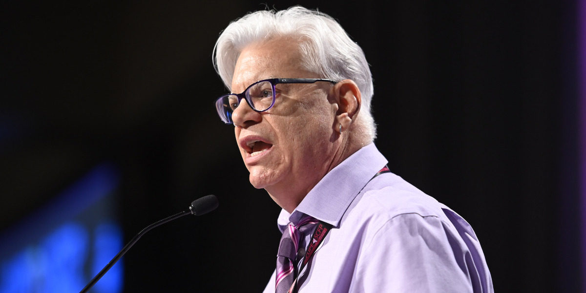 Image shows Fred Hahn speaking at a large event, against a mostly black background, and into a small podium microphone.
