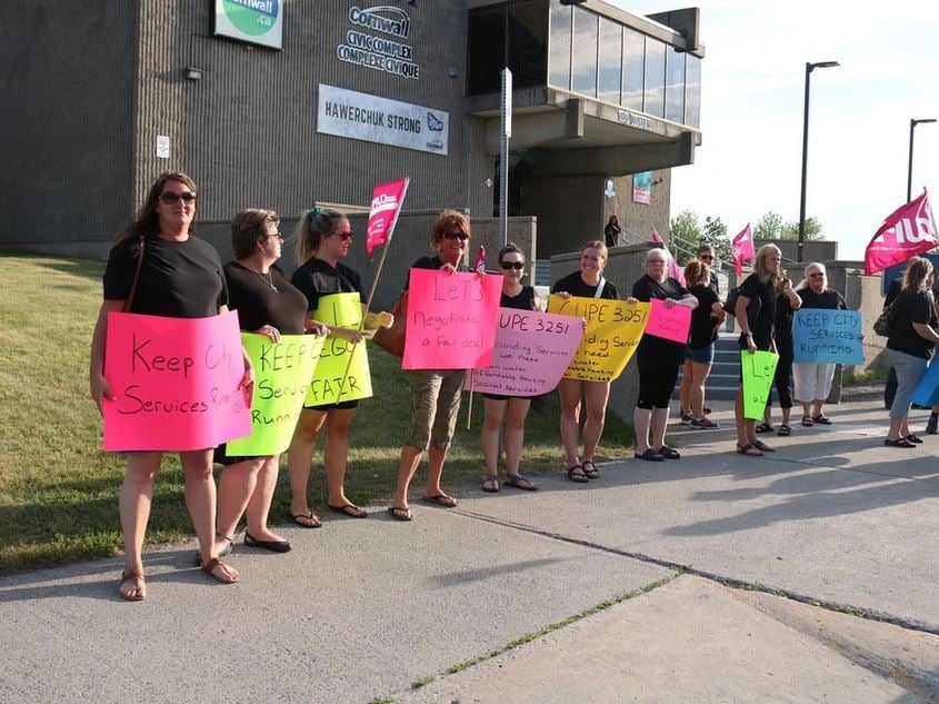Image shows a line of CUPE 3251 workers, holding handwritten signs and pink CUPE Ontario flags, standing outside 