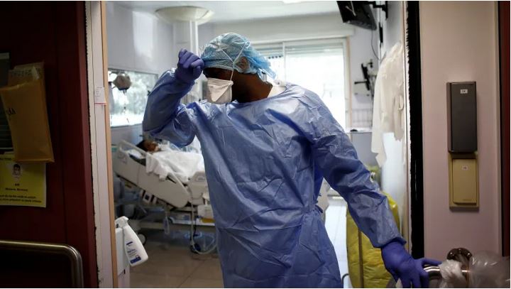 Image shows a nurse in a hospital with their right hand to their forehead, looking to their right, with their torso squarely facing the camera, and in full PPE. Behind them we see a hospital bed. The image feels chaotic.