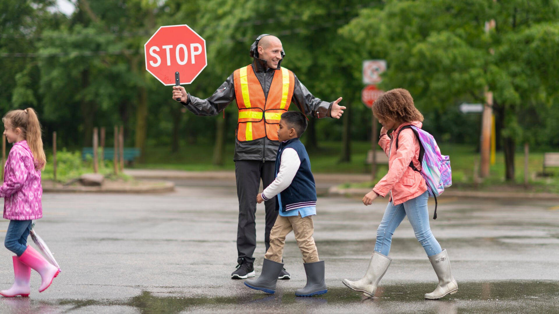A multi-ethnic group of elementary students are on their way to school. The children are crossing the street with the assistance of a smiling crossing guard. The male crossing guard is happy to help the kids stay safe. It's a rainy day and the children are wearing rain coats and rain boots. The crossing guard is holding a stop sign and his wearing a reflective safety vest.